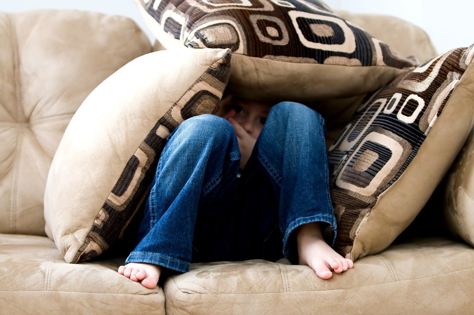 young boy in blue jeans hiding under a pile of cushions on a couch